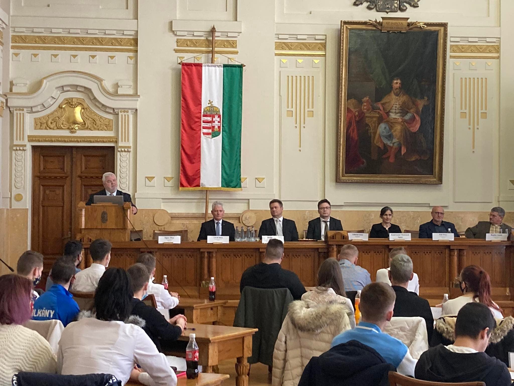 Konferenciaterem, ahol emberek ülnek, és egy hangszóró a pódiumon. egy nagy portré mellett magyar zászló lóg, a résztvevők pedig egy hivatalos rendezvényen jelennek meg.
