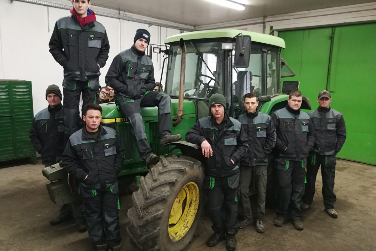 Hét fős csapat zöld és fekete munkaruhában, zöld traktorral pózolnak egy garázsban. egyesek a traktoron állnak, míg mások elöl.