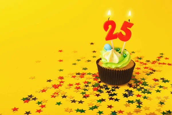 A zöld cukormázzal ellátott cupcake tetején egy 25-ös gyertya világít, élénk sárga háttér előtt. Színes, csillag alakú konfetti van elszórva a cupcake körül, ünnepi hangulatot teremtve. A gyertyák égnek, a 25. évforduló jegyében.