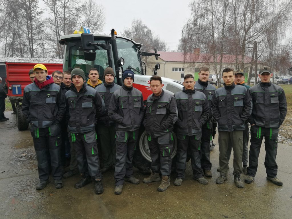 Tizennégy fiatal férfiból álló csoport hozzáillő sötétszürke egyenruhában, zöld díszítéssel, egy nagy traktor előtt állva egy felhős napon.