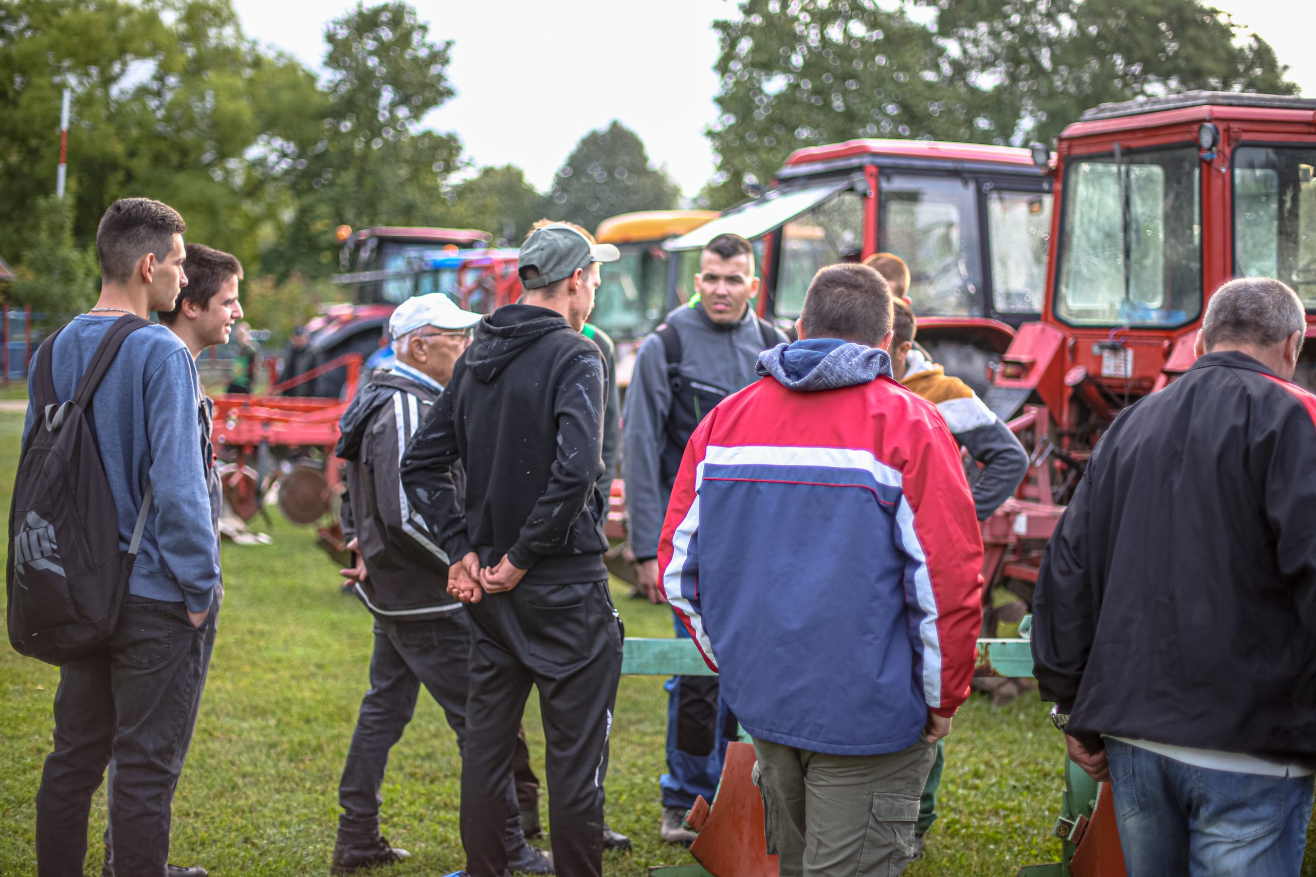 Emberek csoportja, köztük több különböző korú férfi, egy füves mezőn állva, régi piros traktorok között beszélgetnek egy szabadtéri rendezvényen.