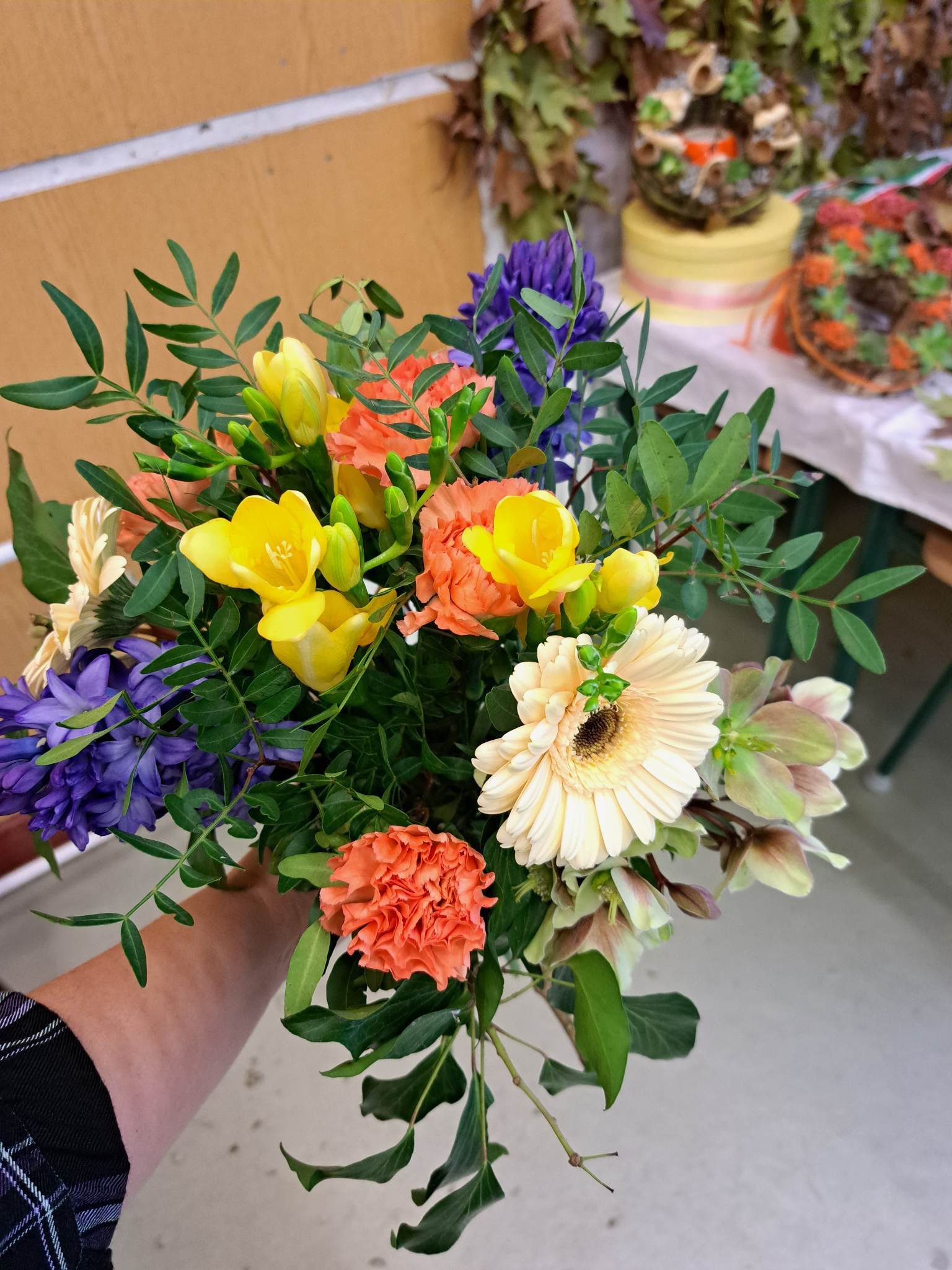 Egy kéz egy élénk csokrot tartva sárga fréziákkal, narancssárga rózsákkal, lila agapantusszal és krémes gerberákkal, válogatott növények hátterében.