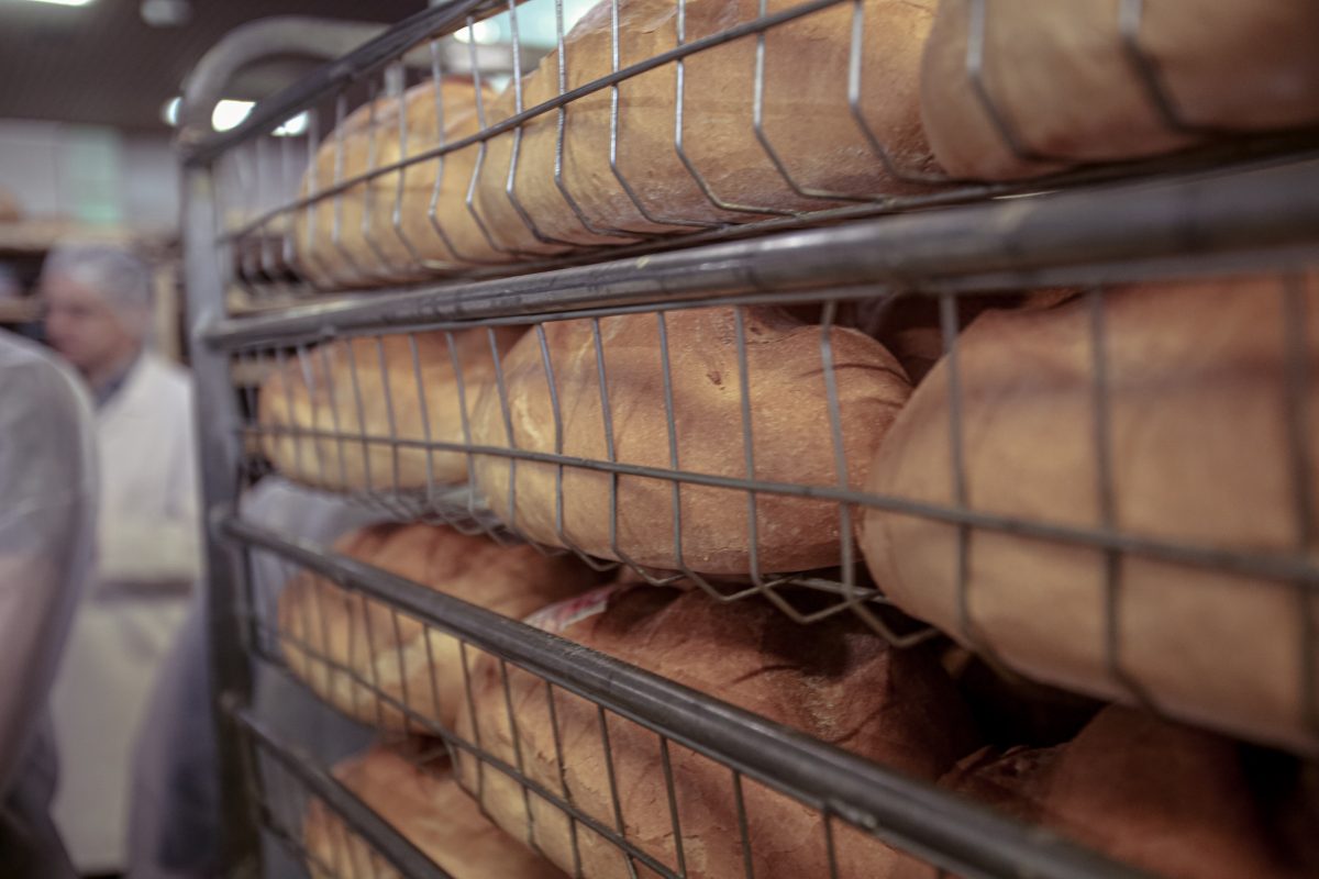 Frissen sült kenyér egy pékség fémpolcaira halmozottan, a háttérben egy fehér egyenruhát viselő, elmosódott pék.
