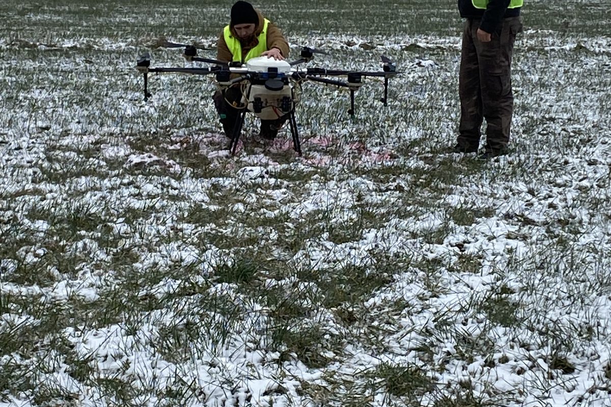 Két ember biztonsági mellényben, egy térdelve, egy nagy drónt vizsgál meg egy havas mezőn. egy személy távirányítót tart, áll. fehér kötegek és csupasz fák a háttérben.