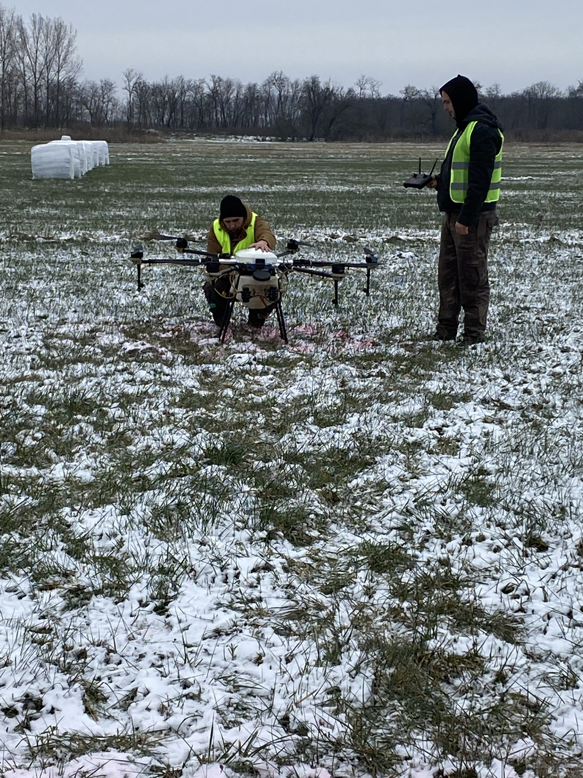 Két ember biztonsági mellényben, egy térdelve, egy nagy drónt vizsgál meg egy havas mezőn. egy személy távirányítót tart, áll. fehér kötegek és csupasz fák a háttérben.