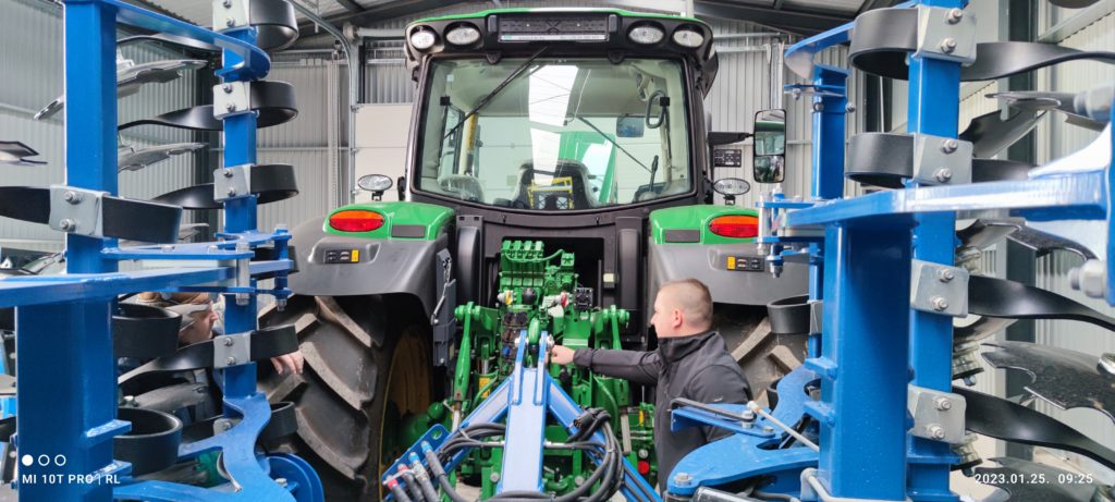 Egy személy egy műhelyben, aki egy zöld traktor hátuljához rögzített mechanizmust állítja be egy jól felszerelt fémkamrában.