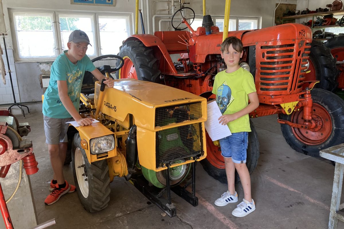 Két fiatal fiú egy garázsban áll régi traktorokkal, az egyik egy sárga traktort érint, miközben a kamerát nézi.