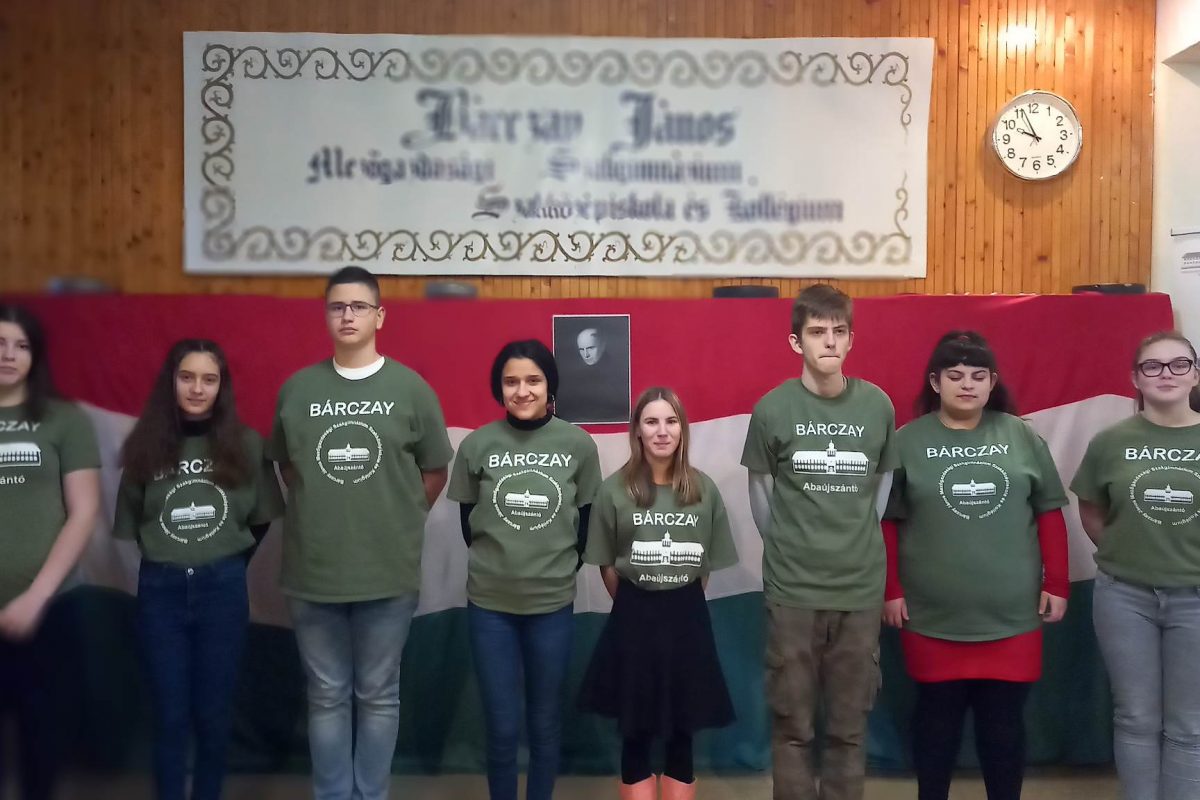 Fiatal felnőttek csoportja, akik "barczay" logóval ellátott zöld pólót viselnek, és egy inspiráló idézettel ellátott transzparens alatt állnak a hallban.