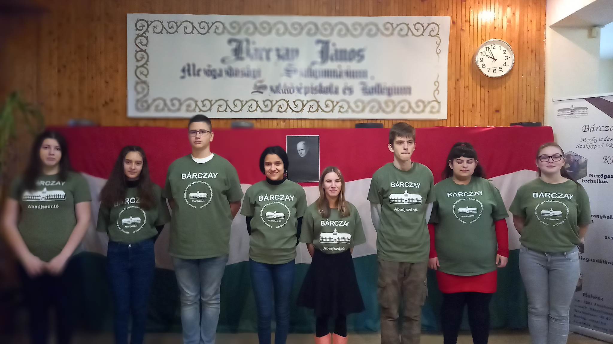 Fiatal felnőttek csoportja, akik "barczay" logóval ellátott zöld pólót viselnek, és egy inspiráló idézettel ellátott transzparens alatt állnak a hallban.