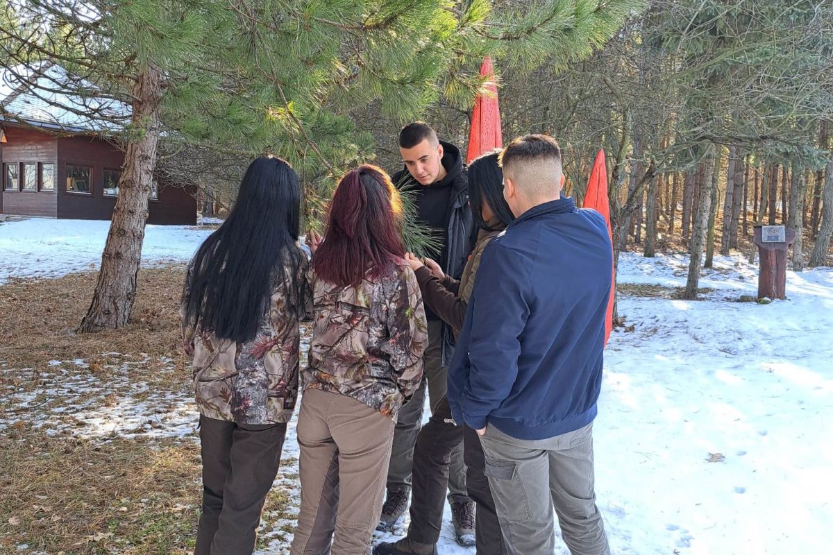 Négy erdőőr egyenruhába öltözött egyén beszélget egy havas, erdős területen, körülötte fenyőfákkal, háttérben egy faépítménnyel.
