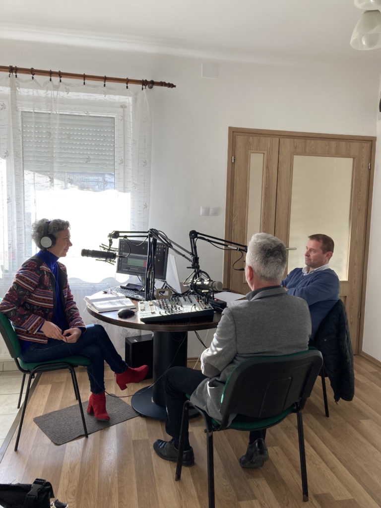 Három ember, két férfi és egy nő, egy jól megvilágított szobában ülve beszélgetnek egy mikrofonnal és felvevőkészülékkel felszerelt podcast-rendszer körül.