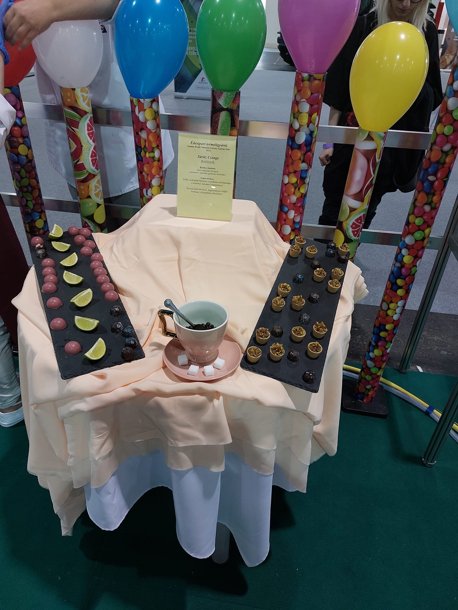 Különféle csokoládékat bemutató kijelző egy elegánsan terített asztalon teáscsészével és tányérokkal, színes cukorkákkal teli oszlopokkal és vibráló léggömbökkel körülvéve.