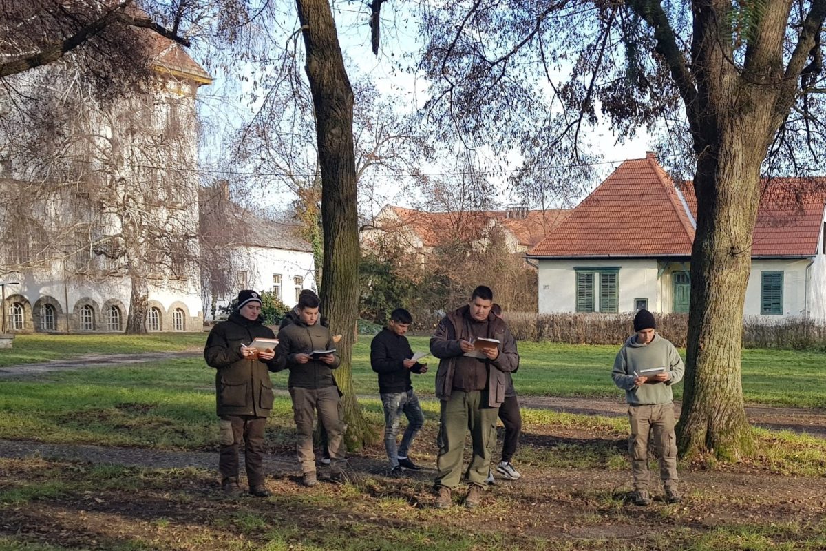 Hat fős, szabadtéri ruházatú, okostelefont használó férfi csoport áll fák közelében, történelmi épülettel és tiszta égbolttal a háttérben.