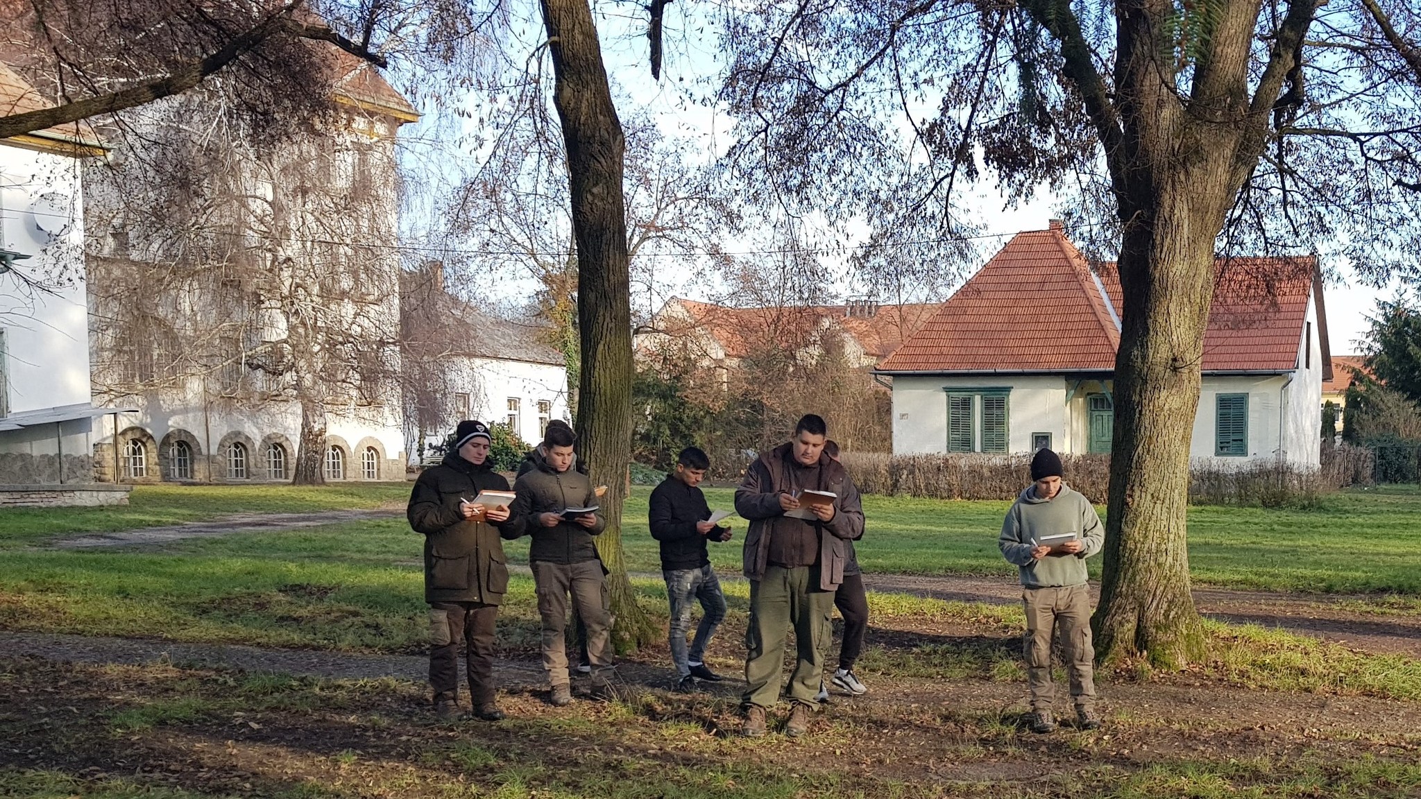 Hat fős, szabadtéri ruházatú, okostelefont használó férfi csoport áll fák közelében, történelmi épülettel és tiszta égbolttal a háttérben.