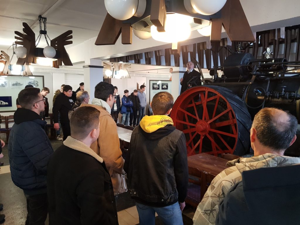 Emberek egy csoportja, akik egy prezentáción vesznek részt egy nagy vintage mozdonykerékkel és egyéb vasúti műtárgyakkal díszített szobában.