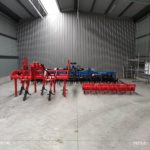 Egy sor mezőgazdasági gép, köztük egy piros kultivátor és egy kék eke, egy tágas, tiszta ipari fészerben tárolva, hullámkarton belsővel.