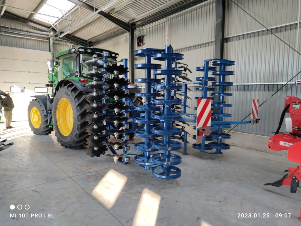 Egy nagy zöld traktor dupla hátsó kerekekkel, egy kék mezőgazdasági tárcsás borona mellett parkolt egy jól megvilágított ipari fészerben, a fotón időbélyeggel, amely 2023 januárjában készült.