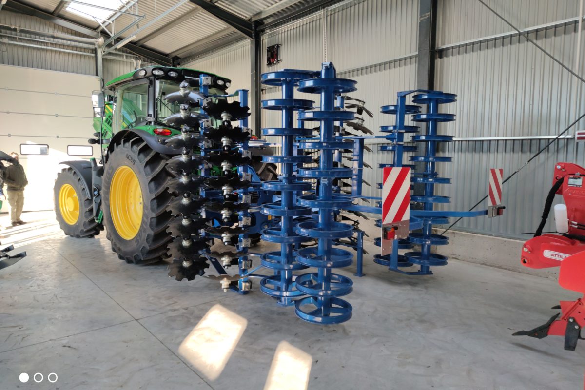 Egy nagy zöld traktor dupla hátsó kerekekkel, egy kék mezőgazdasági tárcsás borona mellett parkolt egy jól megvilágított ipari fészerben, a fotón időbélyeggel, amely 2023 januárjában készült.