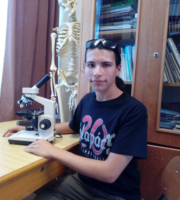 Egy fiatal férfi ül egy mikroszkópos íróasztalnál, mosolyogva, mellette egy csontváz modellel egy könyvekkel teli szobában. fekete pólót és napszemüveget visel.
