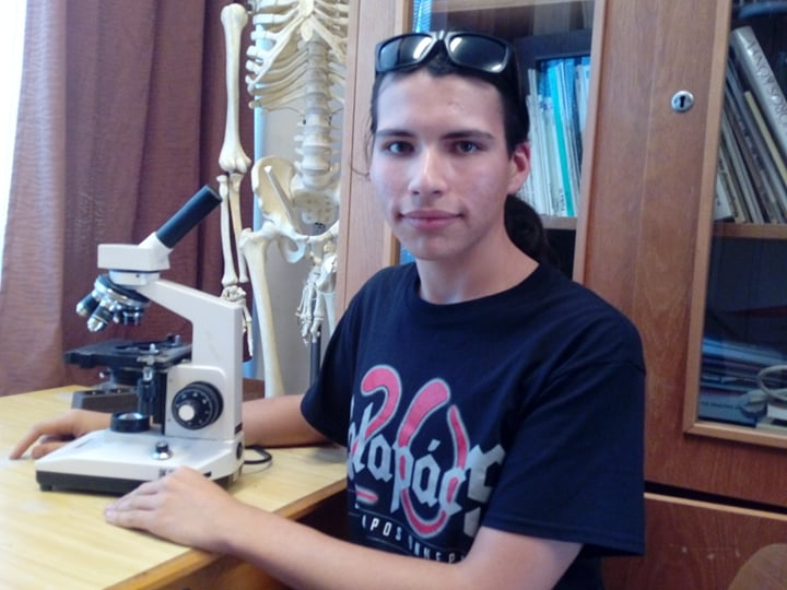 Egy fiatal férfi fekete pólóban és napszemüvegben ül egy asztalnál, mikroszkóppal és egy emberi csontváz modellel a háttérben.