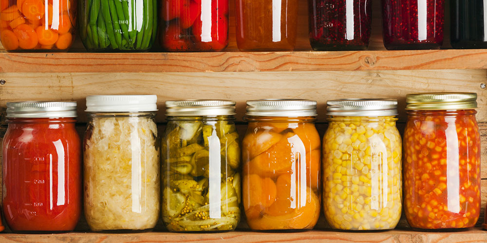 Különféle tartósított zöldségek és gyümölcsök egy sor üvegedényben egy fapolcon, köztük sárgarépa, paprika, uborka, savanyúság, kukorica és paradicsom.
