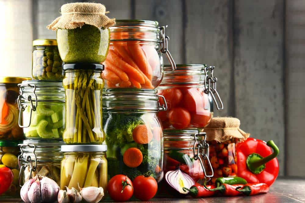 Különféle üvegedények, amelyek tartósított zöldségeket, például sárgarépát, uborkát és paprikát, valamint friss paradicsomot és fokhagymát tartalmaznak, egy fából készült asztalon.