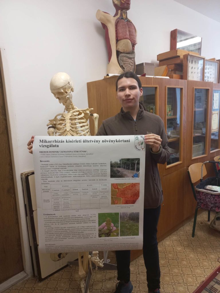 Egy fiatal férfi egy füzetet tartva áll egy emberi csontváz modell mellett egy poszter előtt, amelyen a növénybiológiához kapcsolódó szövegek és képek láthatók, egy szobában, ahol különféle tárgyak és műalkotások találhatók.