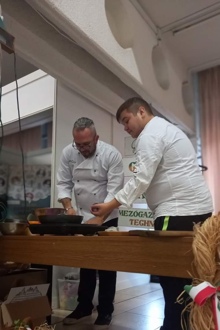 Két férfi szakács, az egyik megfigyelő, együtt főz egy bemutató asztalnál egy oktatási plakátokkal ellátott szobában, egy serpenyőt használva egy hordozható tűzhelyen.