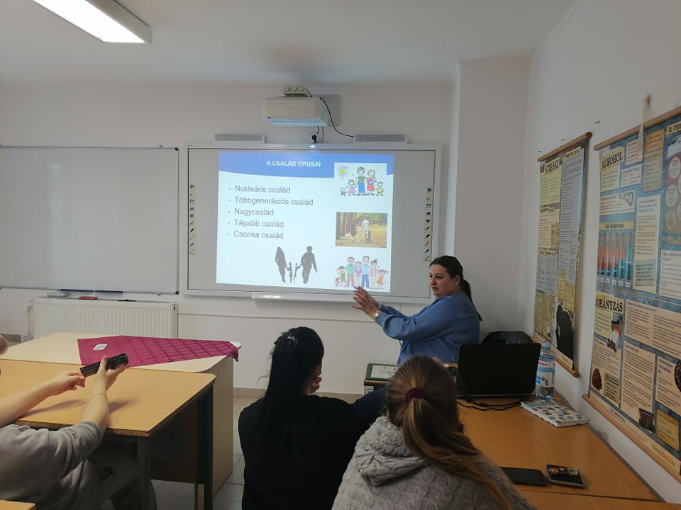 Osztálytermi jelenet egy tanárral, aki egy prezentációs diát magyaráz el a szociális készségekről, képekkel és pontokkal kiemelve az asztalok körül ülő diákoknak.