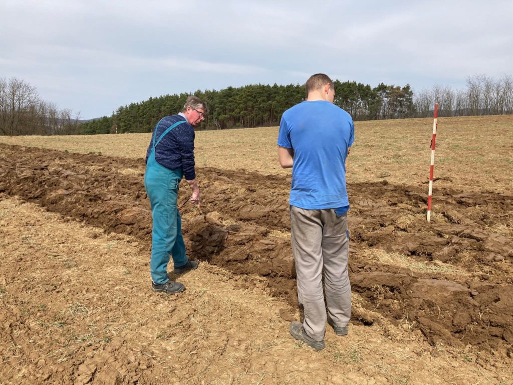Két férfi egy felszántott területen dolgozik, egyik overálban, a másik kék ingben, mérőpálca közelében vizsgálják a talajt.