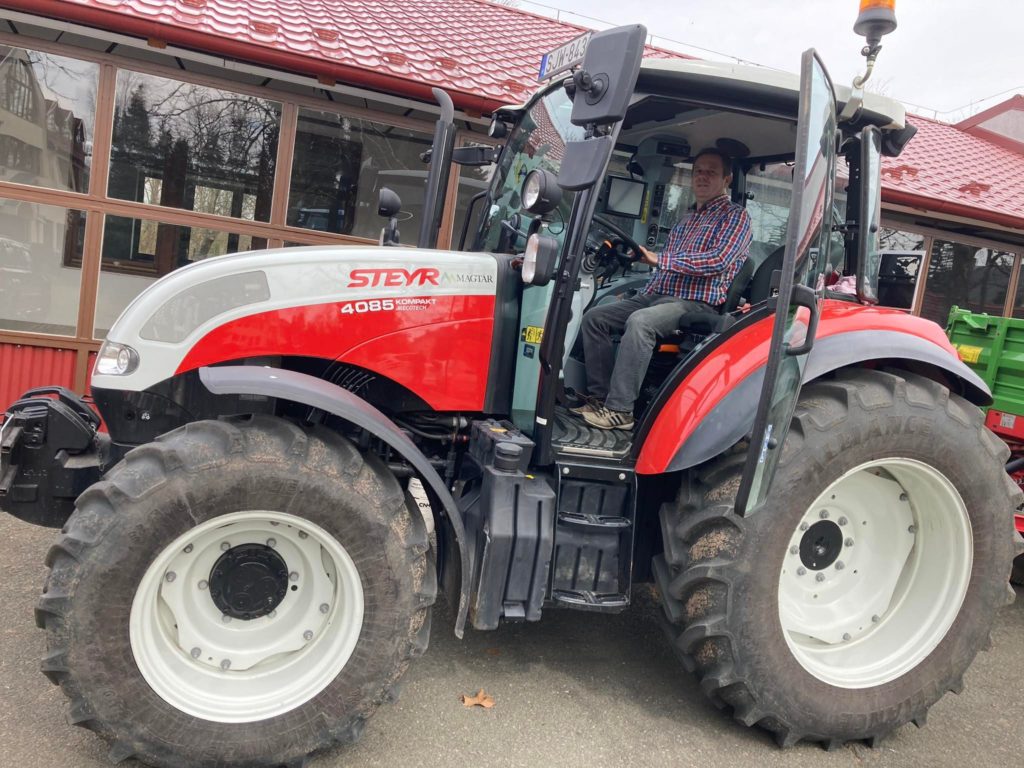 Egy férfi egy nagy, piros-fehér steyr 4085 típusú traktor vezetőülésében ül, a szabadban parkolt egy üvegfalú épület közelében.