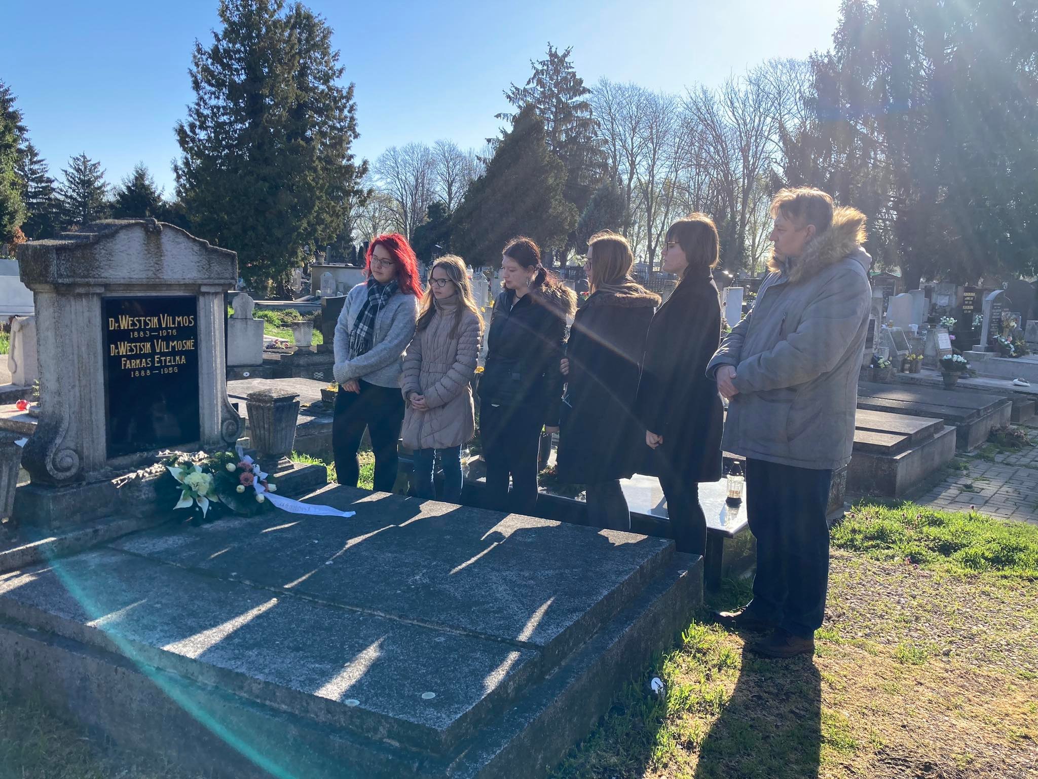 Hat fős csoport áll a virágokkal és sírkővel jelzett sír mellett, a tiszta kék ég alatti temetőben. a jelenet békés, a napfény átszűrődik a fák között.