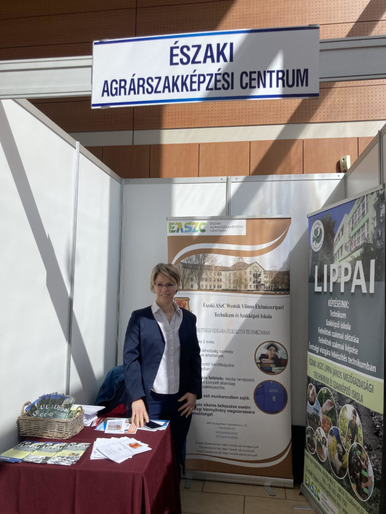 Egy öltönyös nő áll az „északi agrárszakképzési centrum” feliratú stand mellett egy oktatási vásáron, tájékoztató brosúrákkal és plakátokkal.