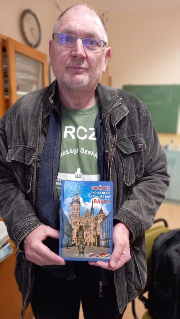Egy középkorú férfi szemüveges, farmerdzsekiben egy grafikus pólón, és egy kassáról szóló könyvet tart maga előtt tantermi környezetben.
