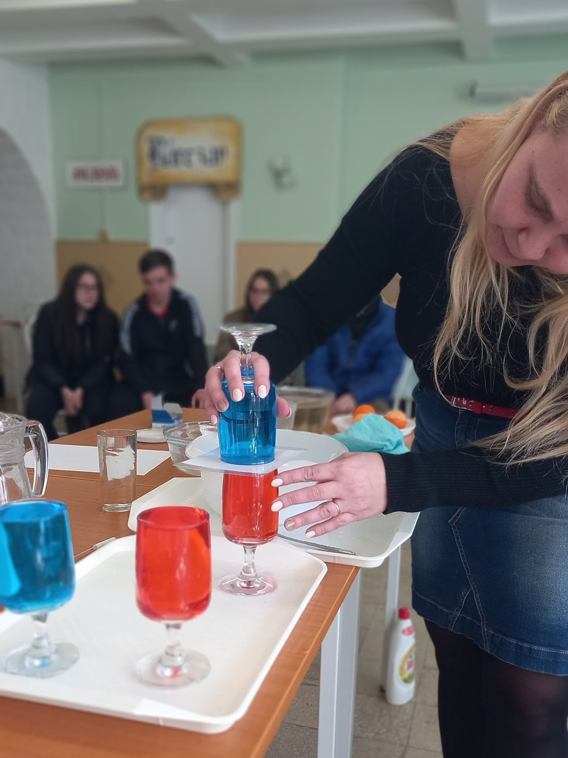 Egy nő tudományos kísérletet végez színes folyadékokkal lombikban, figyelmesen kék folyadékot önt egy lombikba, miközben hallgatóság figyeli a háttérben.