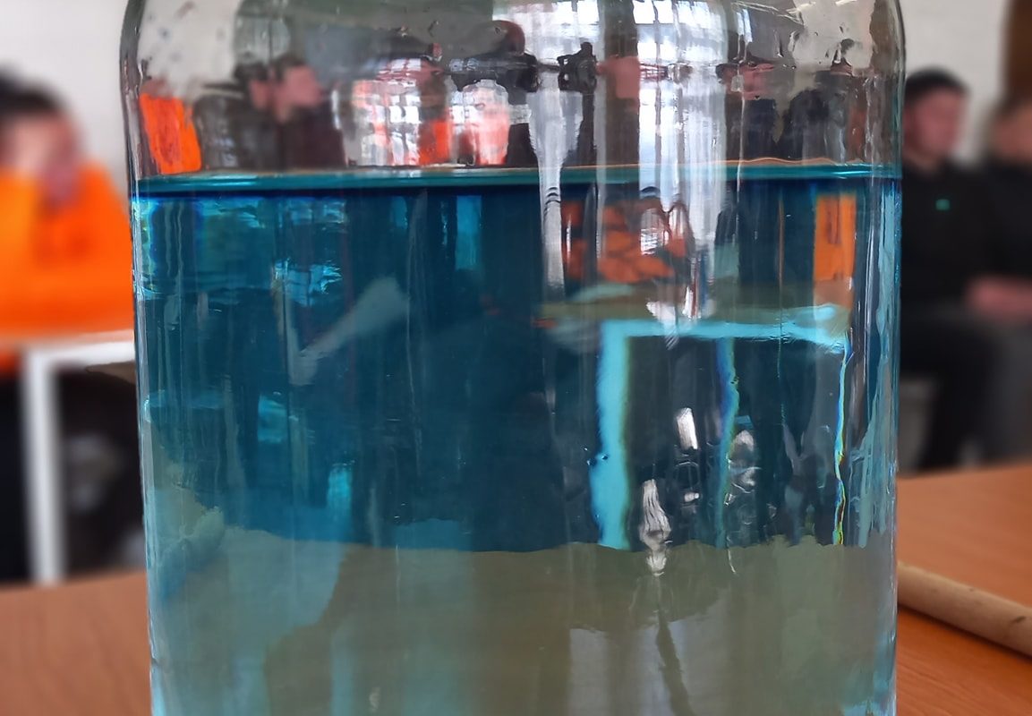 Egy nagy, kék folyadékkal töltött üvegedény az asztalon, a háttérben elmosódott emberekkel, beltéri környezetben.