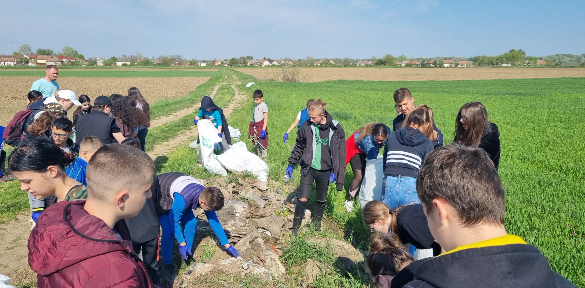 Diákok és felnőttek csoportja, akik egy vidéki zöld mezőn vesznek részt egy takarítási rendezvényen, tiszta kék ég alatt gyűjtik a hulladékot.