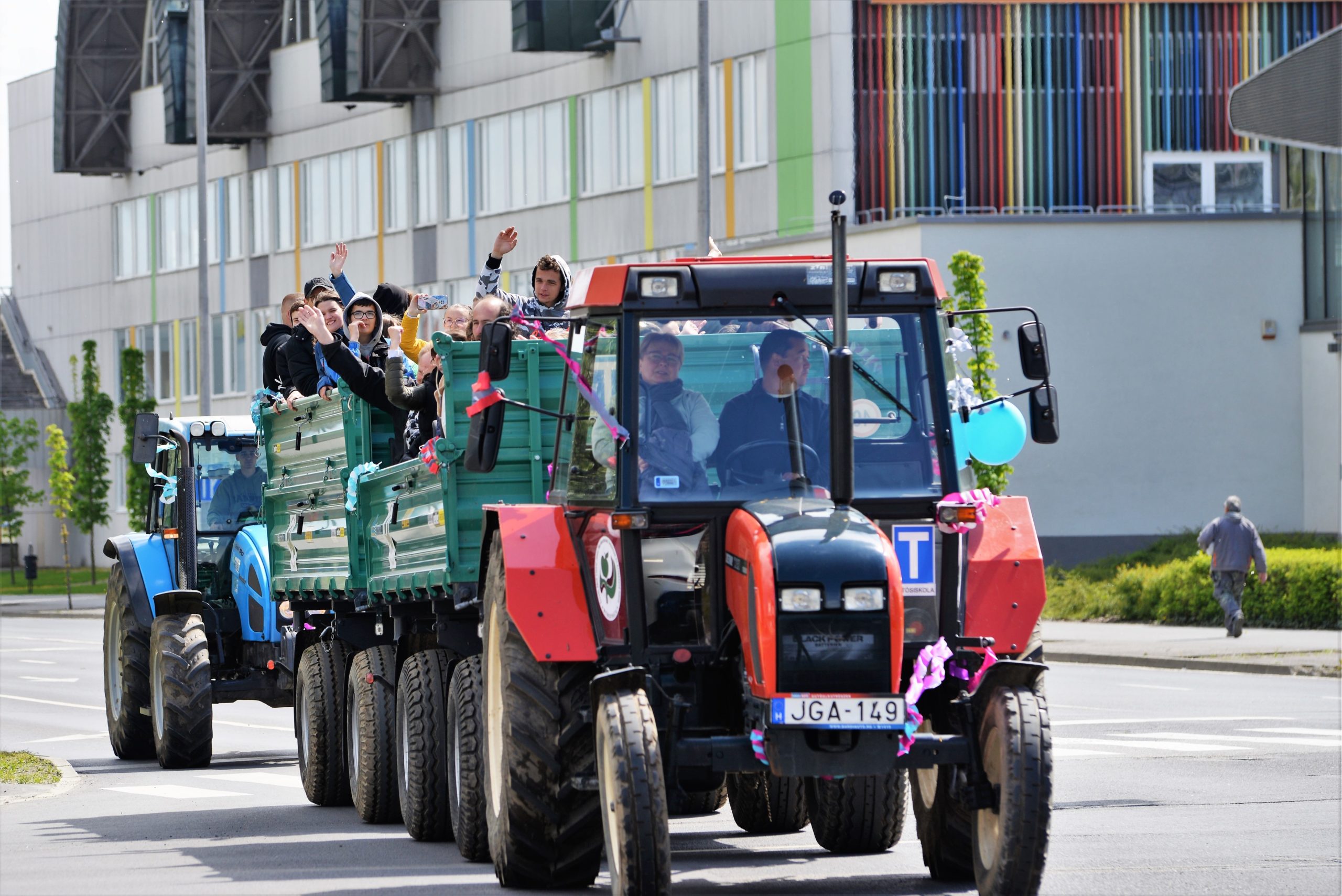 Emberek egy csoportja ujjong az ünnepi díszekkel díszített pótkocsi hátuljáról, amelyet egy piros traktor vontatott egy napsütötte utcán, valószínűleg felvonulás vagy ünneplés közben.