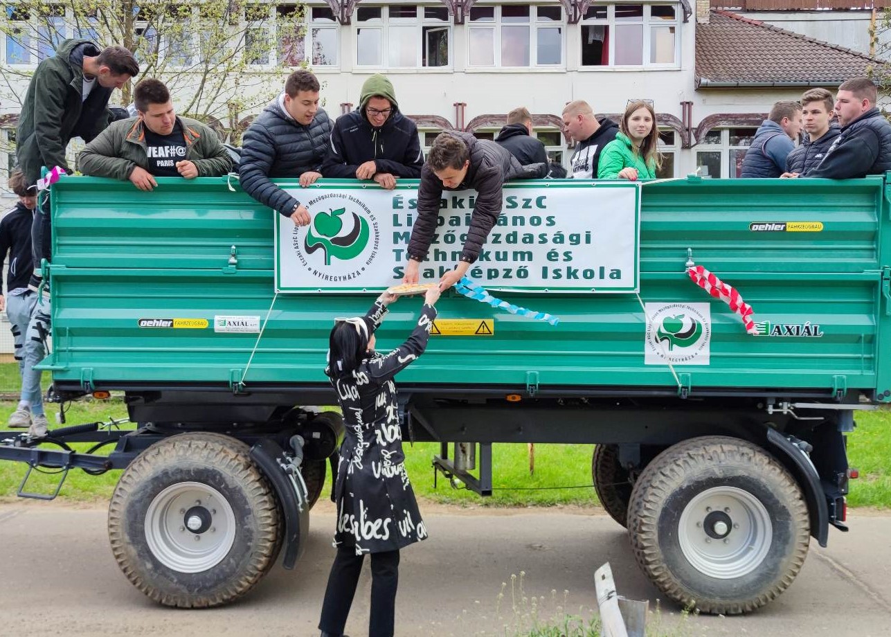 Diákok és tanárok csoportja összegyűlik egy zöld trailer körül, iskolai emblémákkal ellátott transzparensekkel egy szabadtéri rendezvényen. egy fekete-fehér ruhás diáklány interakcióba lép a trailerrel.