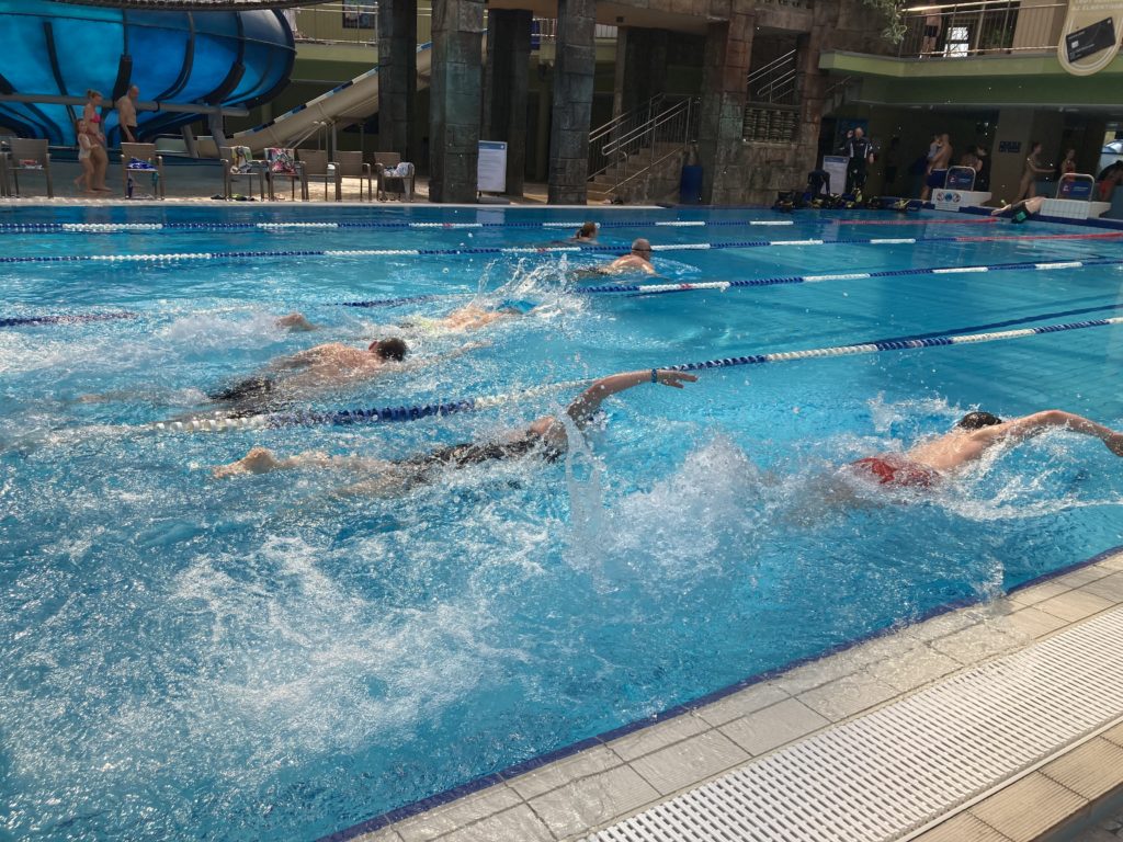 Úszók versenyeznek egy fedett medencében, és fröccsenést keltenek a szabadfogású ütés végrehajtása közben, a háttérben nézőkkel.