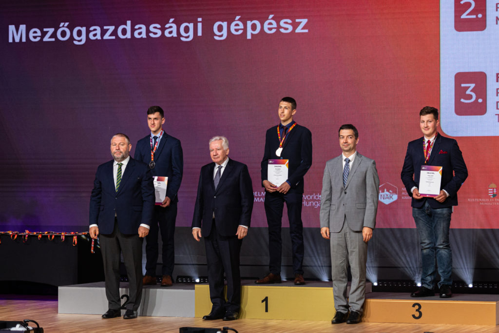 Öt férfi egy díjátadó színpadon, oklevelekkel, érmekkel, háttérben egy nagy képernyővel, amelyen a rangsor és a "mezőgazdasági gépész" cím látható.