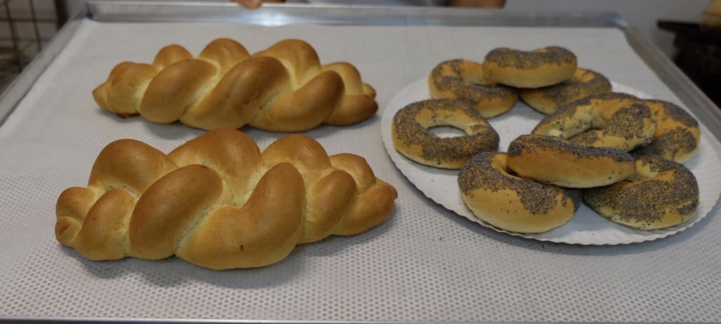 Frissen sült kenyér tálcán: bal oldalon csavart cipók, jobb oldalon mákos bejgli, mindez pékségben látható.