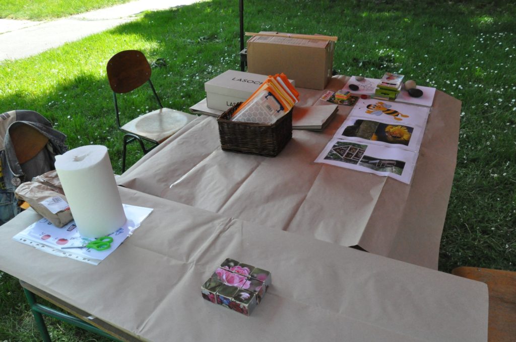 A bézs színű terítővel borított kültéri asztalon különféle tárgyak láthatók, köztük dobozok, kosarak, prospektusok és néhány nyomat természetes fényben, zöld fűvel körülvéve.
