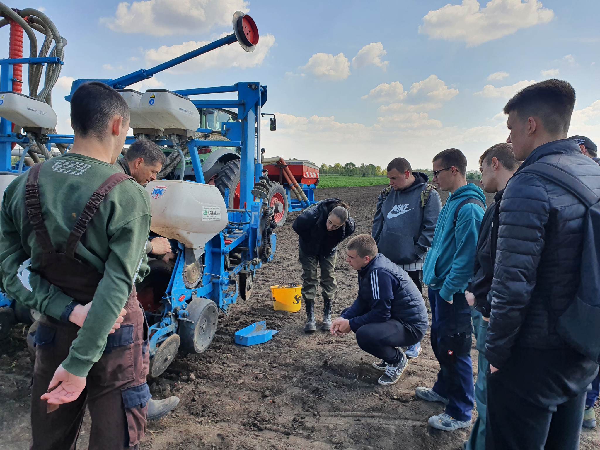 Férfiak egy csoportja mezőgazdasági gépek bemutatóját figyeli egy szántóföldön, és valakire összpontosítva, aki elmagyarázza a berendezés működését.