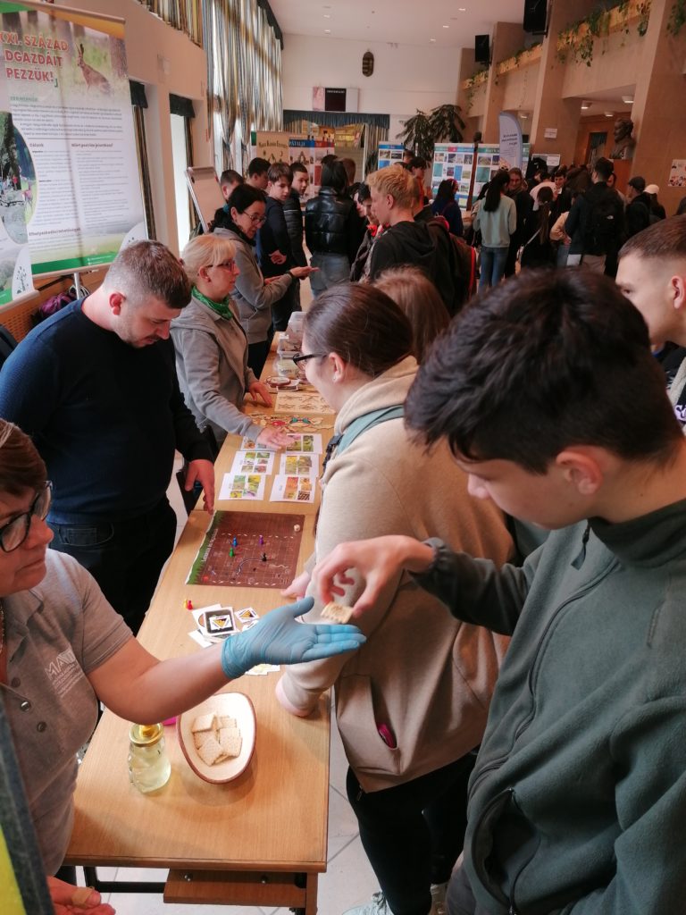 Az emberek egy zsúfolt beltéri rendezvényen vesznek részt, ahol egy információs asztal és interaktív társasjátékok láthatók, interaktív és oktató légkört teremtve.