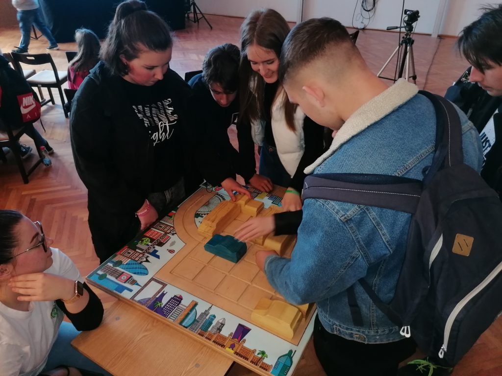 Tinédzserek csoportja, akik társasjátékban vesznek részt, alaposan megvizsgálják és megvitatják a játékelemeket egy asztalon, egy osztálytermi környezetben, a háttérben felállított kamerával.