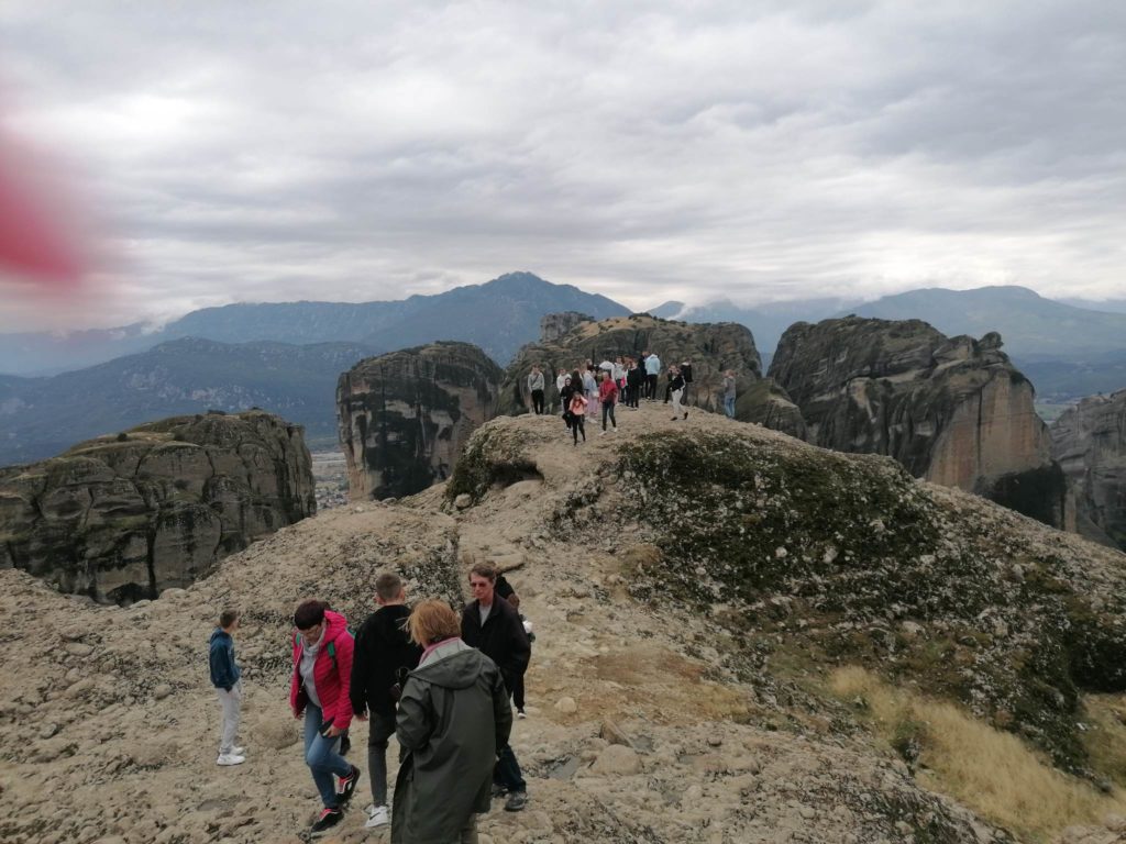 Egy csoport ember áll és sétál egy sziklás gerincen, drámai sziklaképződmények és hegyek a háttérben a felhős ég alatt. A táj zord és kiterjedt, kaland és felfedezés érzését keltve.