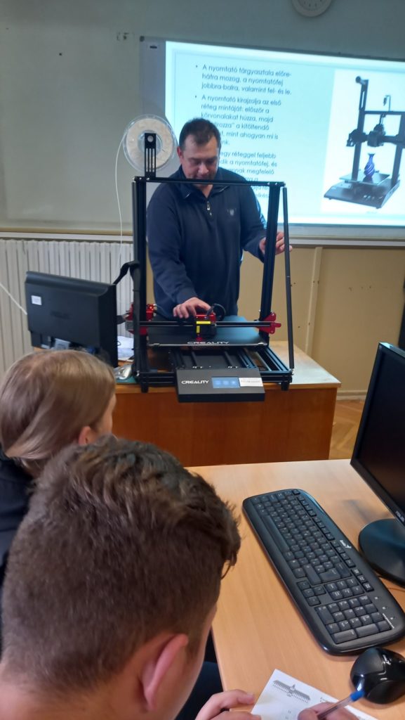 Egy tanár az osztályterem előtt áll, és három diáknak mutat be egy 3D nyomtatót. Mindannyian egy asztal körül vannak számítógép-monitorokkal, billentyűzetekkel és papírmunkával. A tanár mögötti képernyőn megjelenik egy prezentációs dia a 3D nyomtatóról.