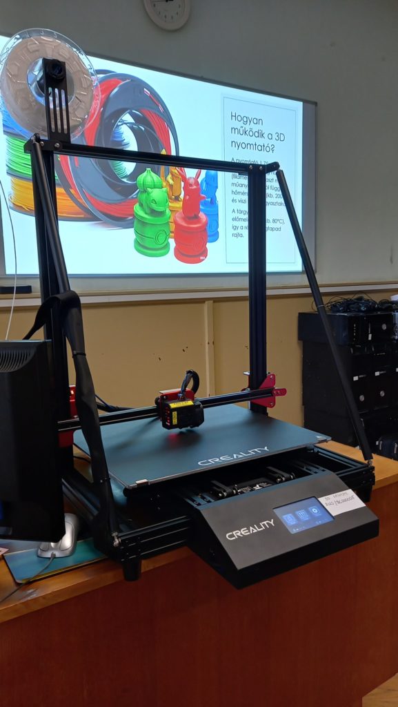 Az osztályteremben egy asztalon ül egy Creality 3D nyomtató, mellette egy monitoron színes 3D-nyomtatott sakkfigurák és magyar szöveg látható a 3D nyomtatást magyarázva. A nyomtató be van állítva és használatra kész, a háttérben az izzószálak láthatók.