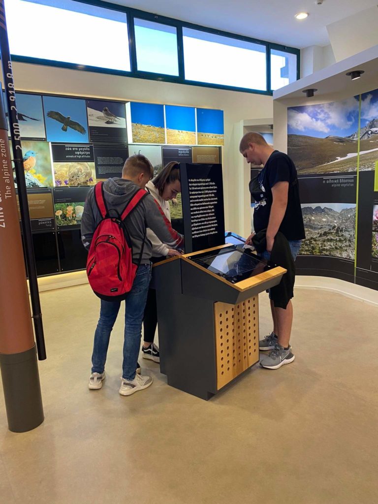 Három ember áll egy interaktív kiállítás körül egy múzeumban. Közülük ketten hátizsákot viselők a képernyőre fókuszálnak, míg a harmadik személy a közelben áll. A falakat informatív táblák és természetfotók szegélyezik.